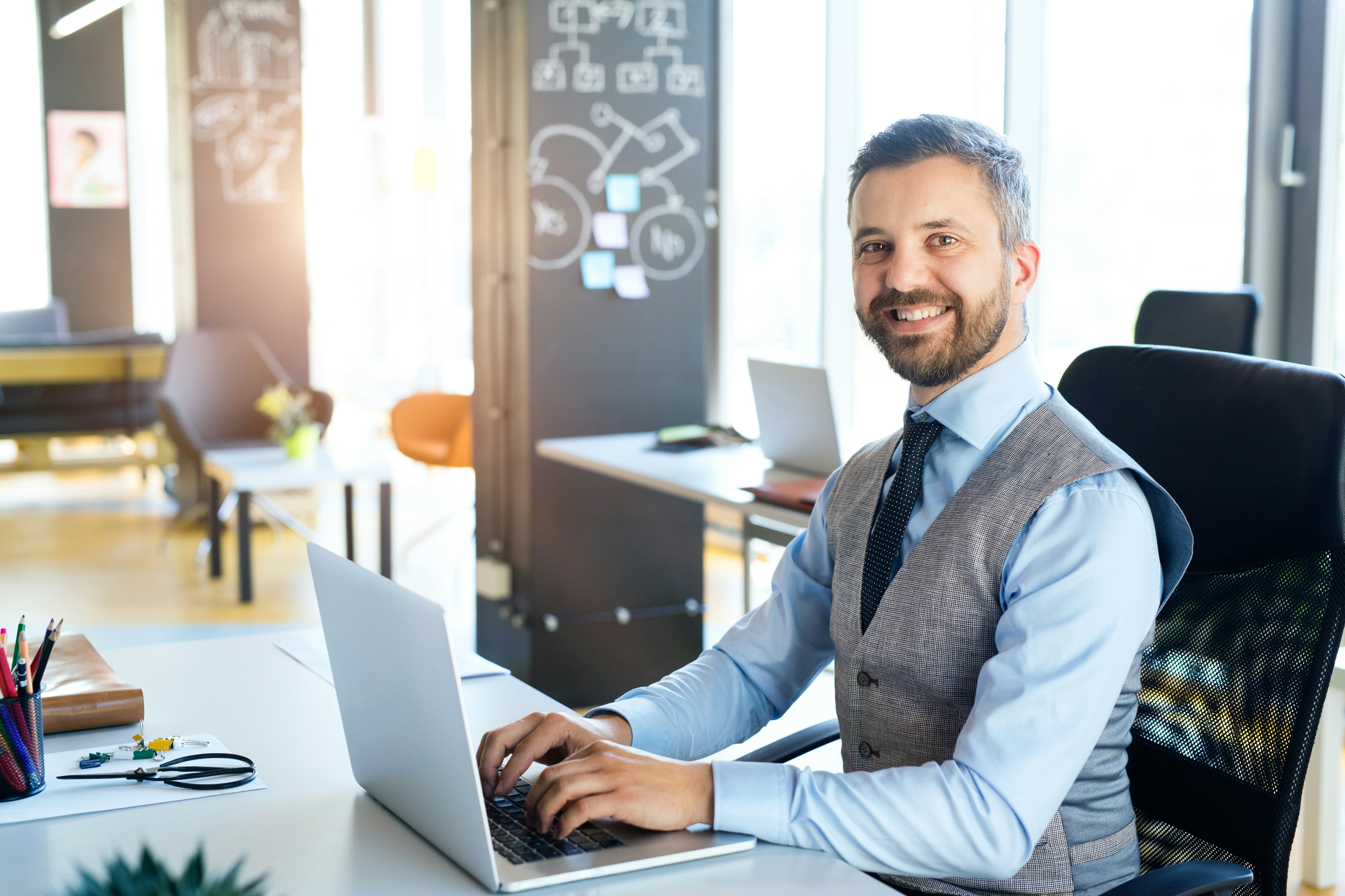 A consultant in a waistcoat and tie working on a laptop in a bright office, with diagrams on a blackboard behind him