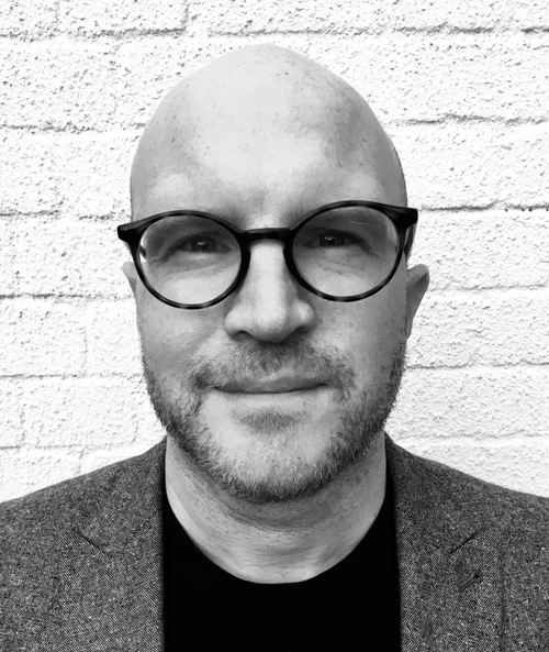 Oliver Hurrey, in black and white, wearing round glasses