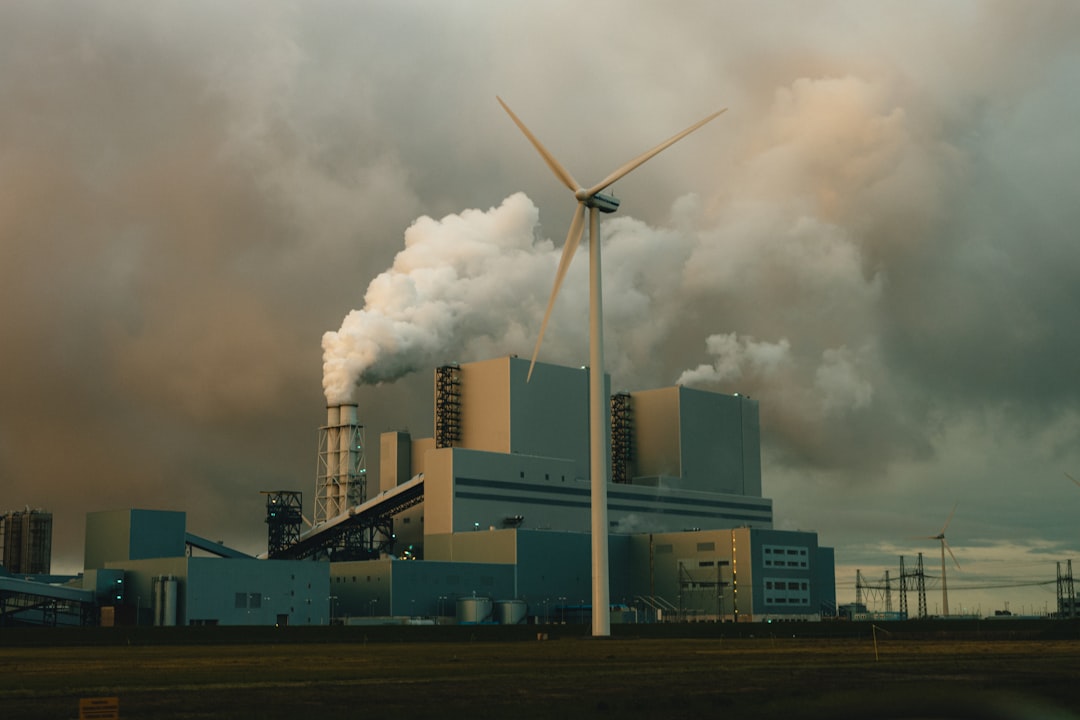 Power station emitting smoke beside a wind turbine under a hazy sky