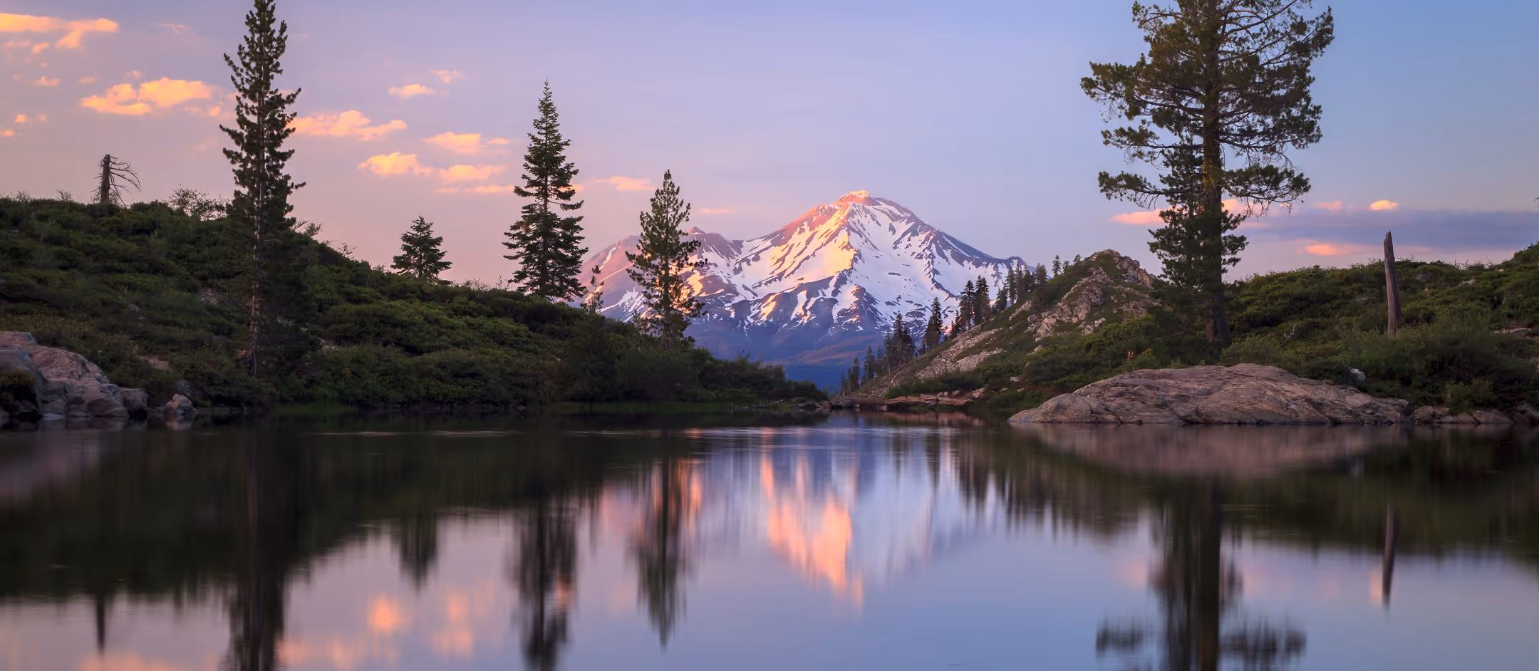 Snow-capped mountain at sunset with trees and rocks reflected in a calm lake.