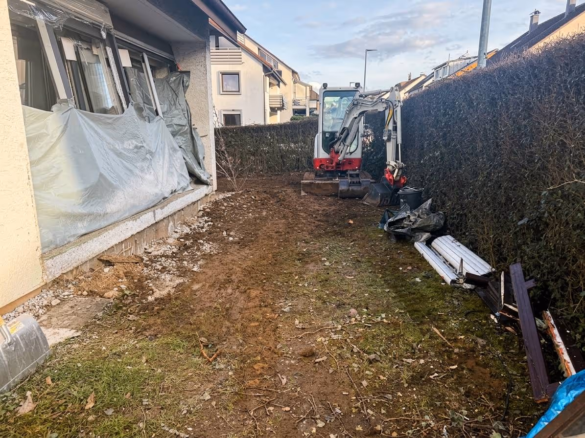 Mini excavator parked in a cleared backyard next to a house with removed windows covered by plastic.