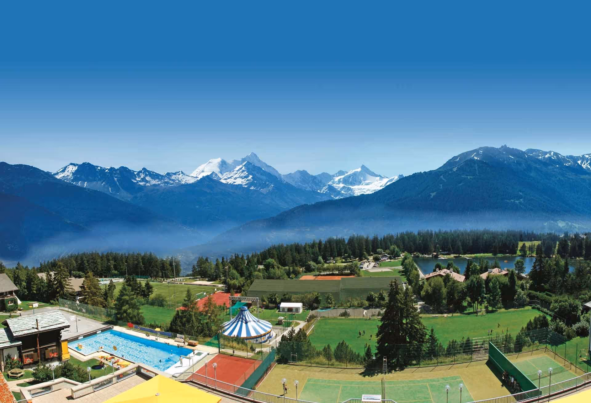 Pool, tennis courts, green fields, forest, and snow-capped mountains under a clear blue sky.