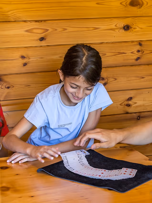 Girl watching a card trick performed on a table with wooden walls in the background.