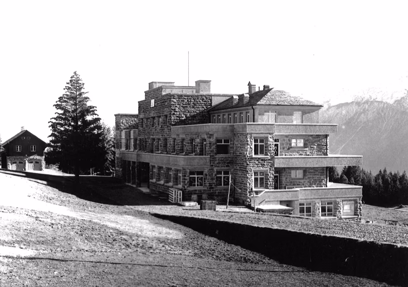 Black and white photo of a large stone building with balconies in a mountainous area.