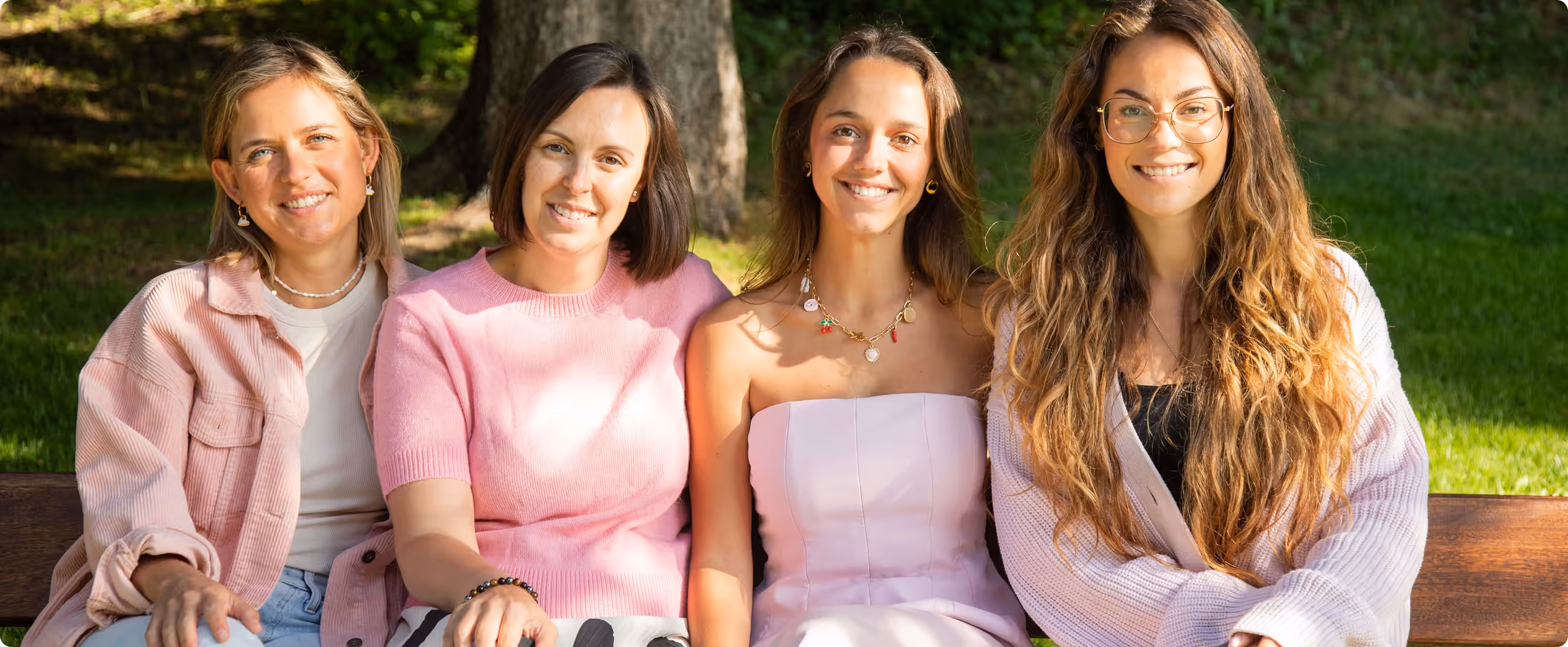 Four smiling women sitting closely on a park bench with trees and grass in the background.