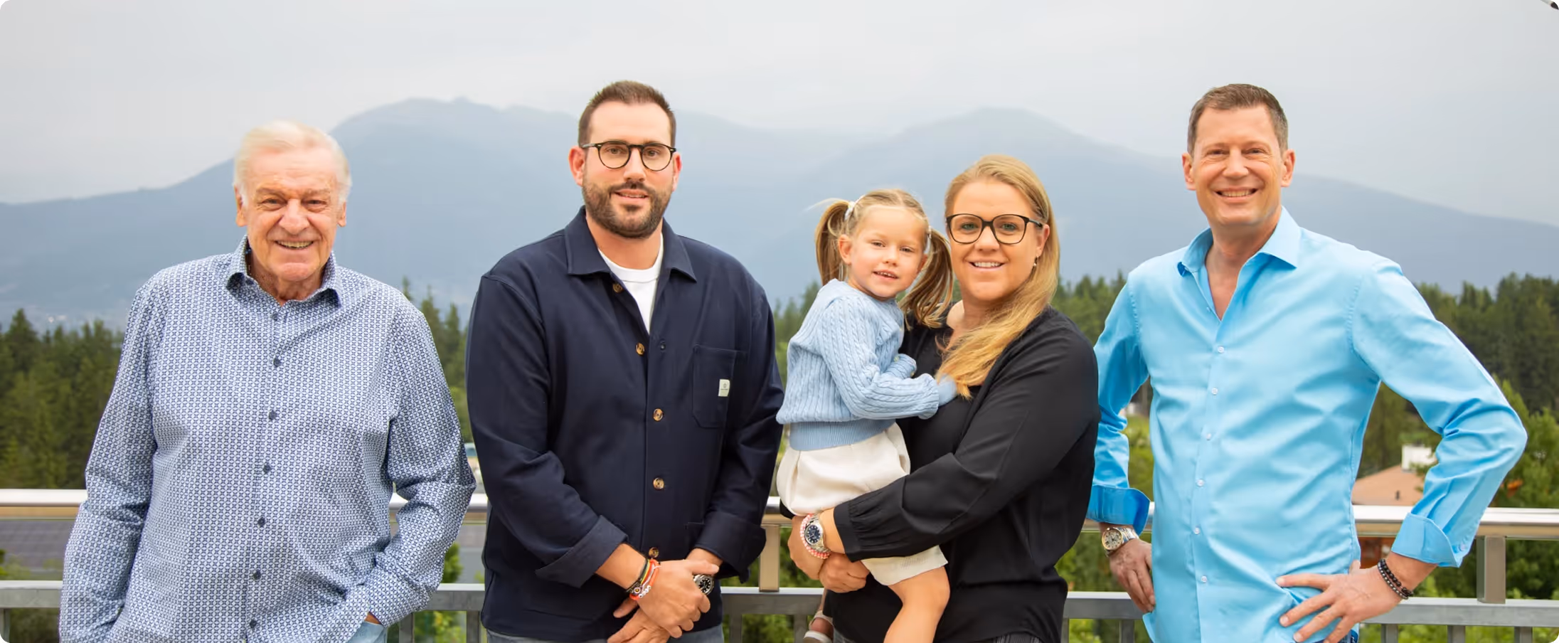 Five people standing on a balcony with mountains and trees in the background, including a child in arms.
