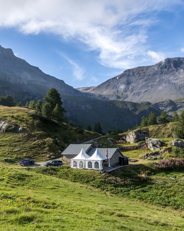 Stone house with white tents and parked cars in a green mountainous landscape under blue sky.