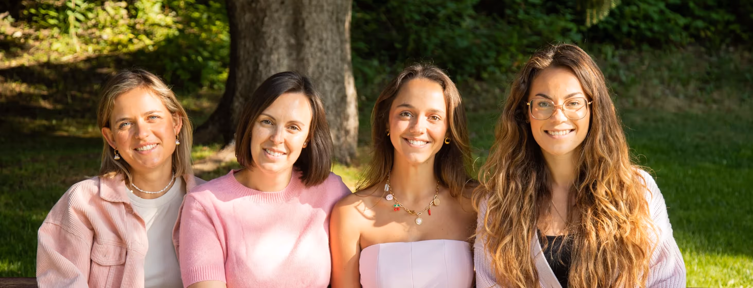 Four smiling women sitting closely on a park bench with trees and grass in the background.