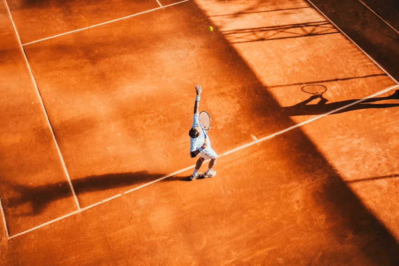 A male tennis player in light clothes serving the ball on a clay court with shadows cast nearby.
