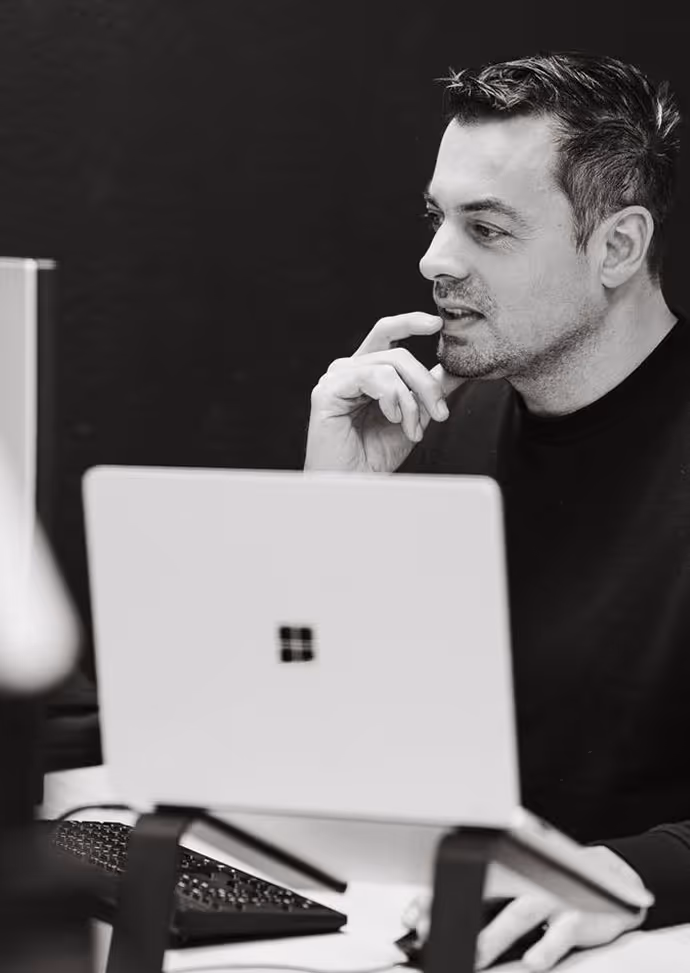 Man die nadenkt terwijl hij naar een laptop scherm kijkt, zittend aan een bureau.