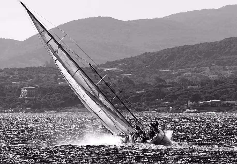 Voilier incliné sur l'eau avec équipage naviguant sur une mer agitée devant des collines boisées.