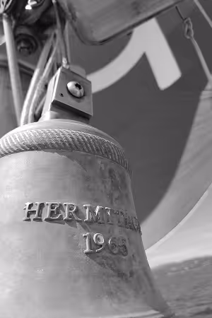 Cloche métallique gravée 'Hermitage 1963' suspendue avec cordelette, en arrière-plan un voile de bateau.