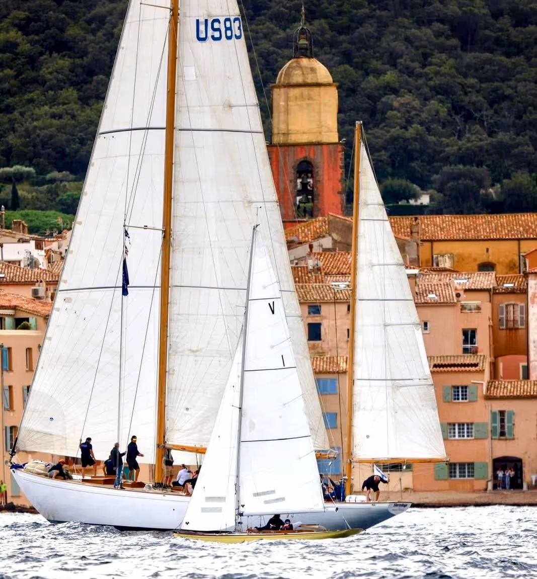 Deux voiliers naviguant sur une eau calme avec un village coloré et une tour cloche en arrière-plan.