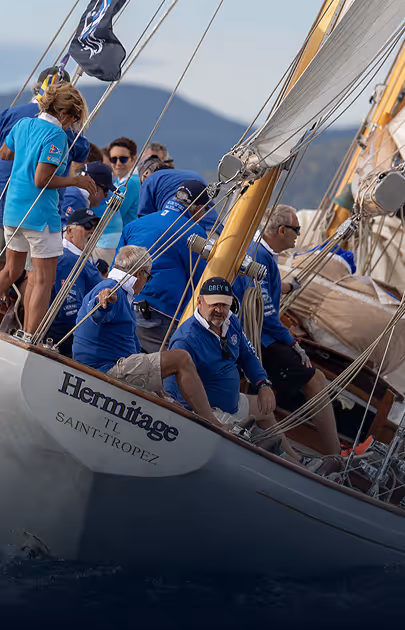 Équipage de plusieurs personnes en vêtements bleus naviguant sur un voilier nommé Hermitage à Saint-Tropez lors d'une régate.