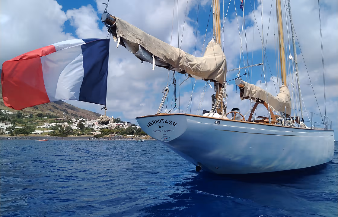 Voilier nommé Hermitage à Saint-Tropez avec un drapeau français flottant et une côte avec des maisons en arrière-plan.