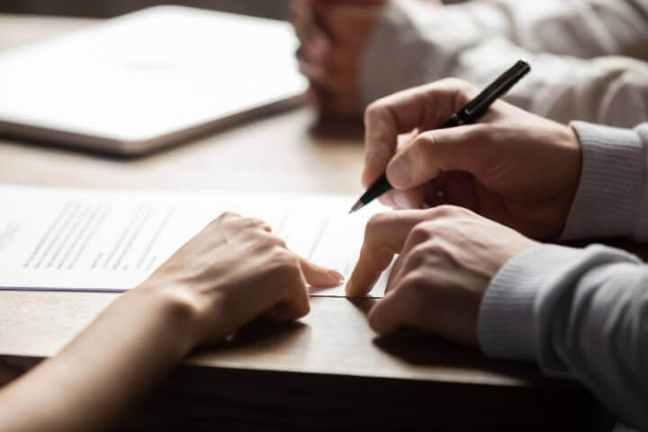 Person zeigt mit dem Finger auf ein Dokument, während eine andere Person mit einem Stift unterschreibt.