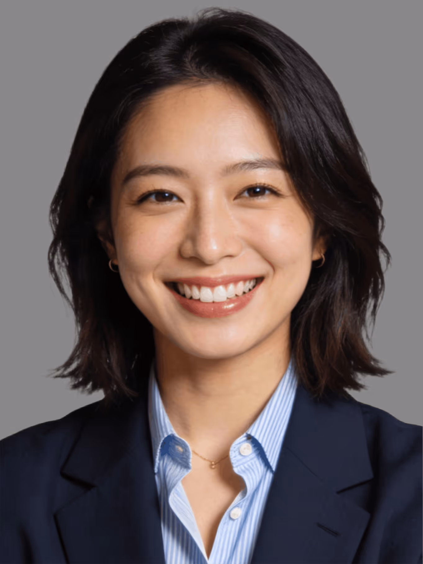 Smiling woman with dark hair wearing a navy blazer and light blue striped shirt against a gray background.