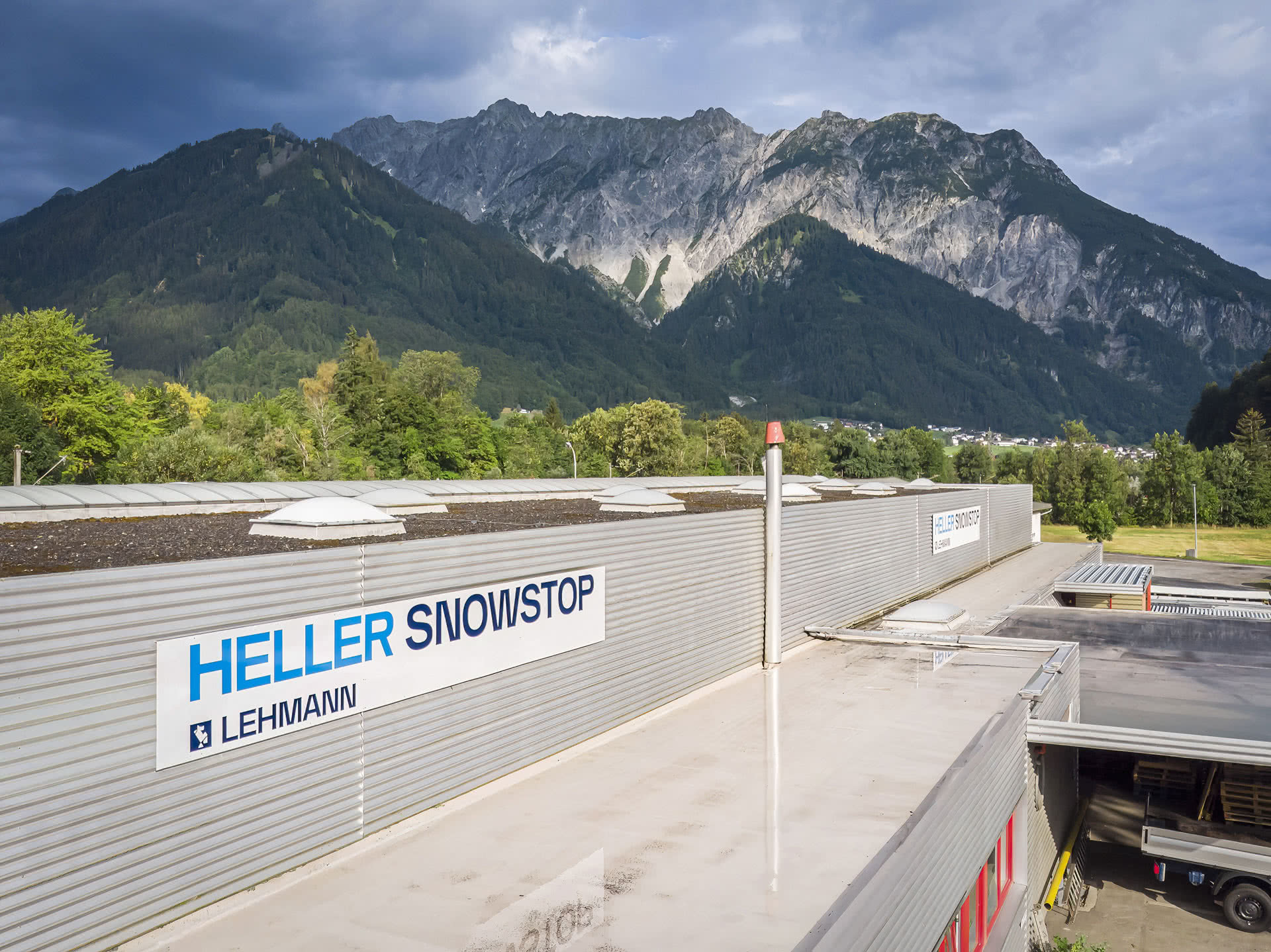 Industriegebäude mit HELLER SNOWSTOP Beschilderung vor grünen Bergen unter bewölktem Himmel.