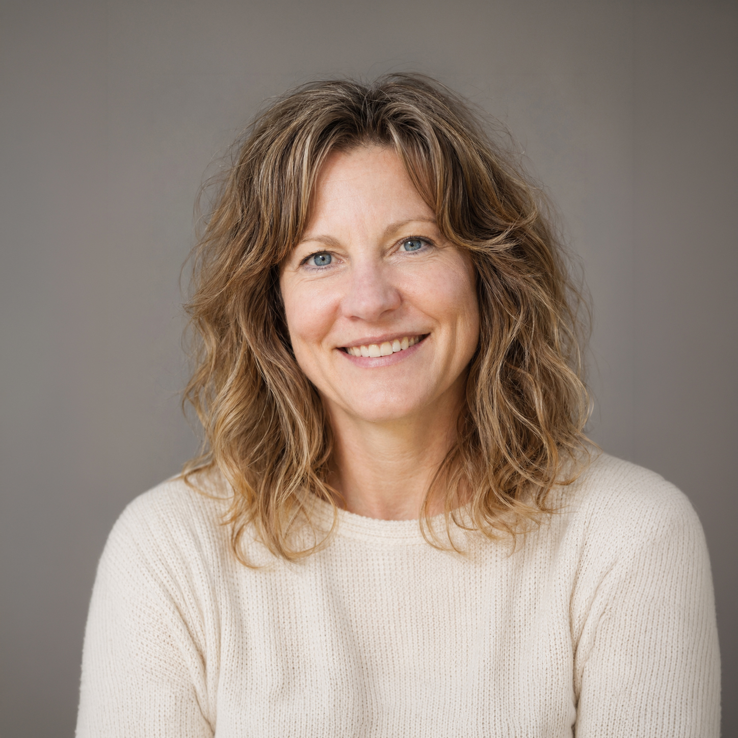 Smiling middle-aged woman with wavy light brown hair wearing a cream sweater against a neutral background.