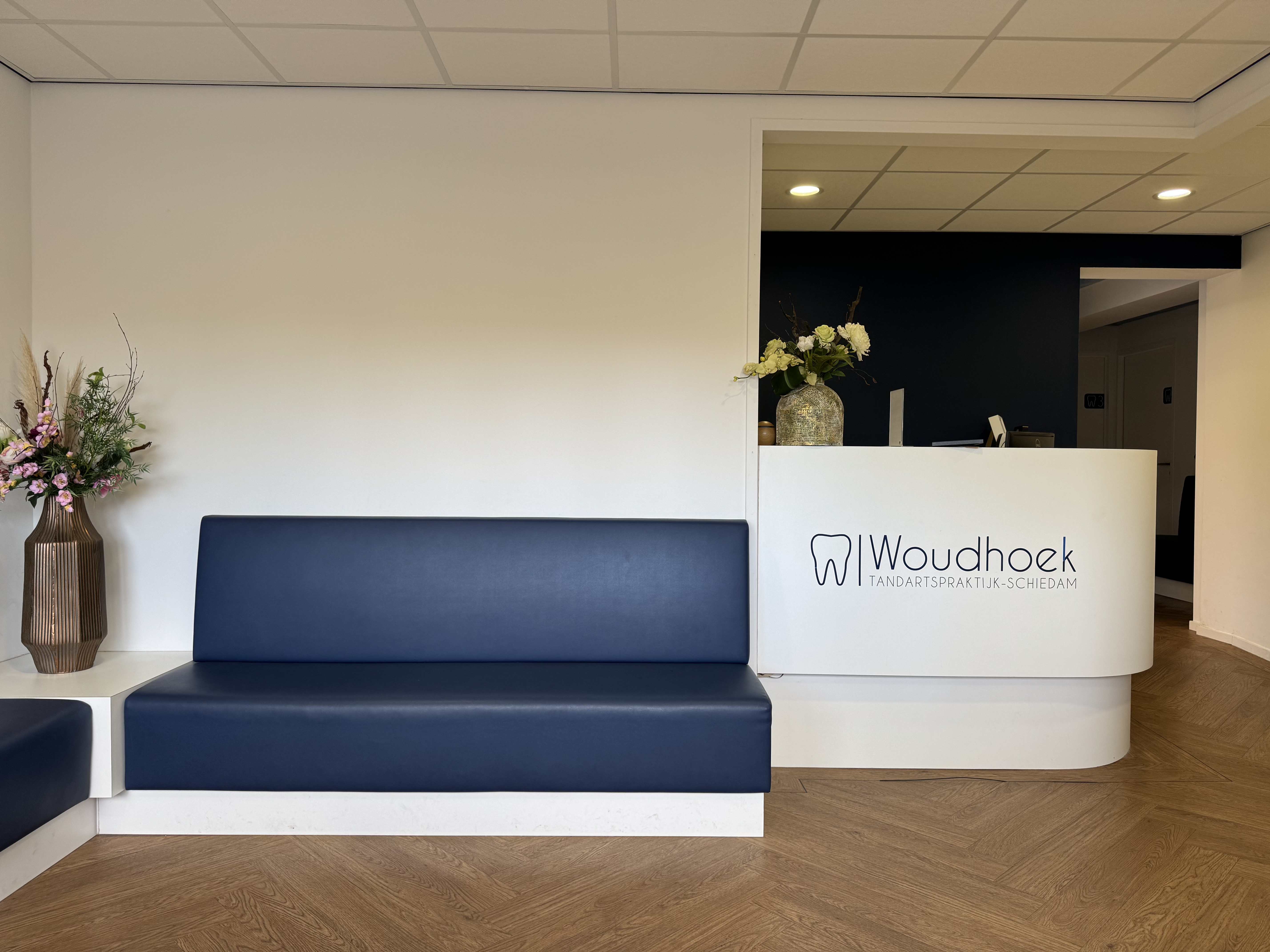 Wachtkamer met blauwe banken, bloemen en balie van tandartspraktijk Woudhoek Schiedam.