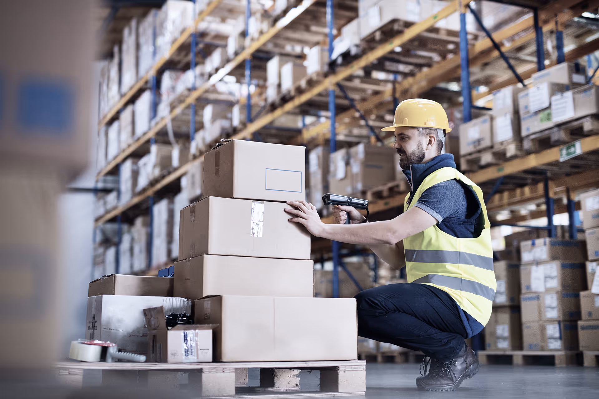 Warehouse worker in yellow safety vest and hard hat scanning boxes stacked on a pallet with a handheld barcode scanner.