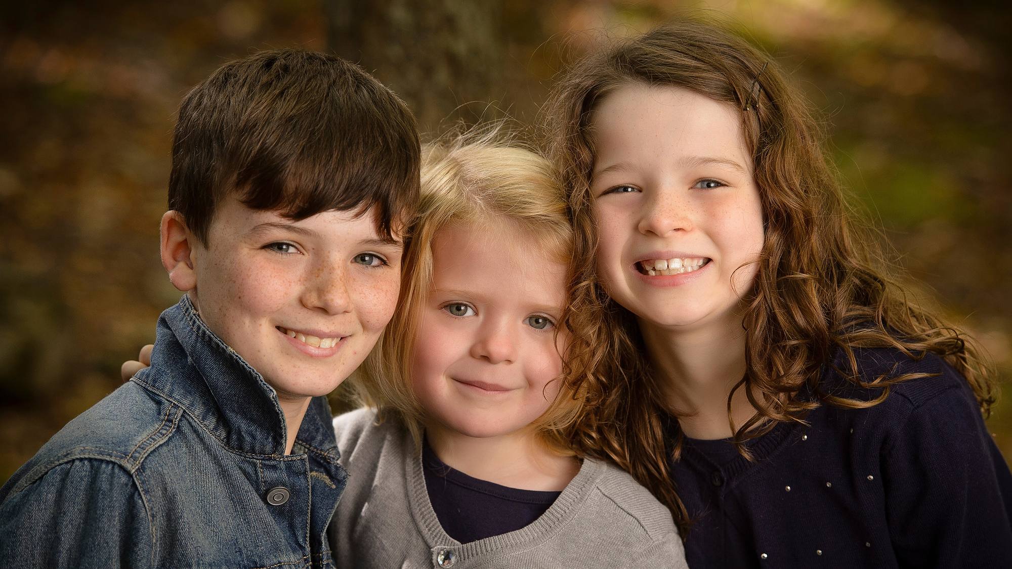 Three young siblings close together against an autumn-toned studio backdrop