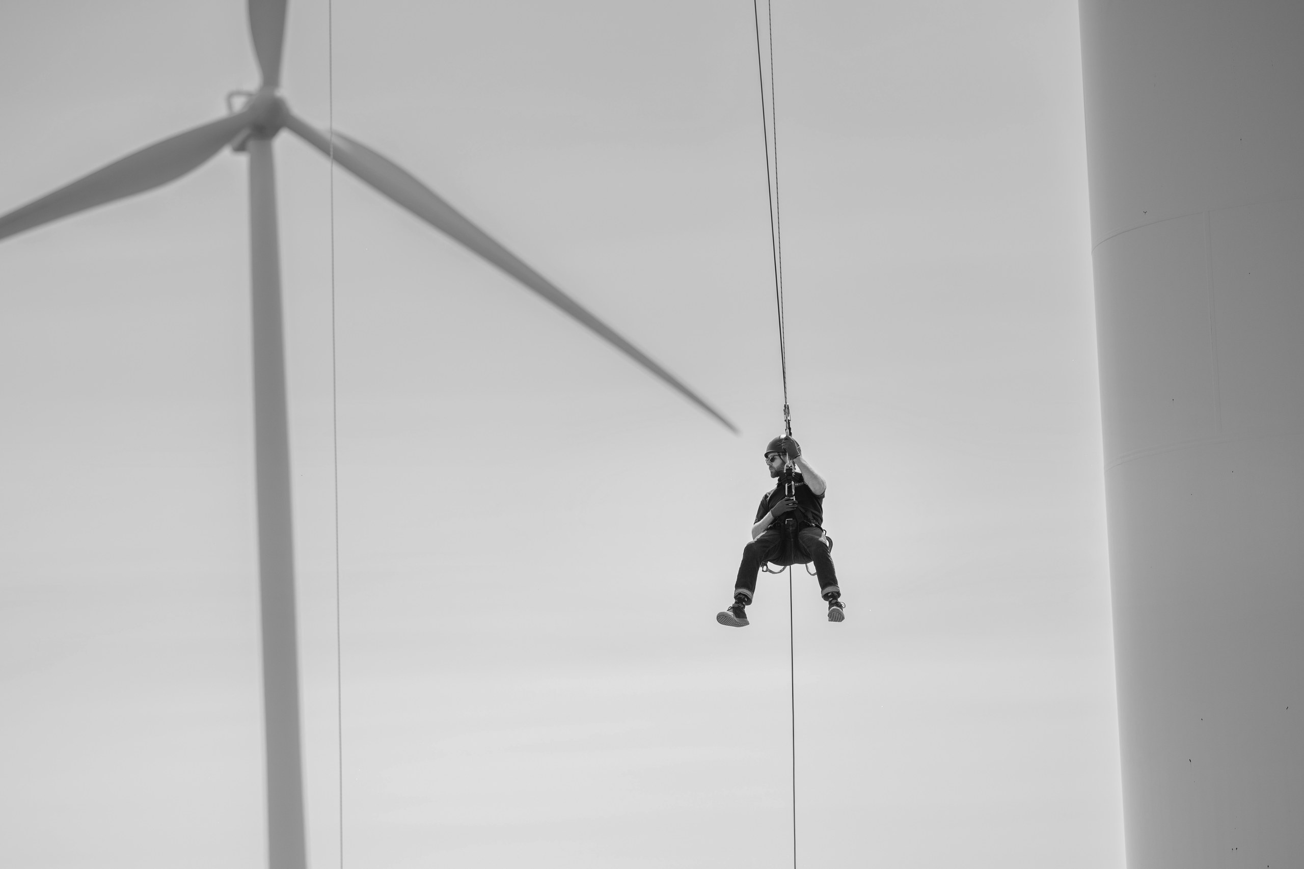 Black and white photo of a rope access technician suspended on lines beside a wind turbine tower, Nova Scotia industrial photography