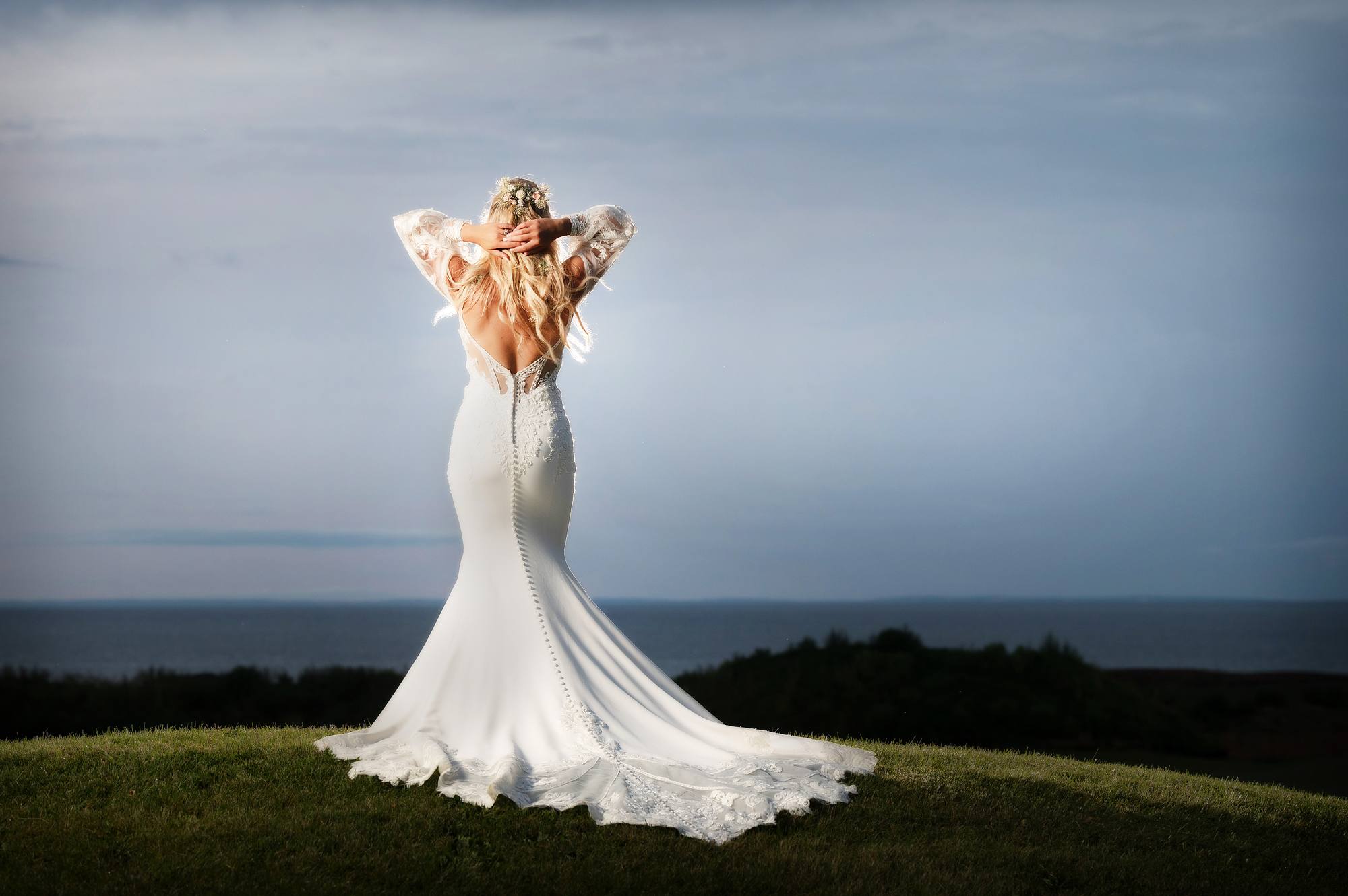 Bride showing the open lace back of her gown on a grassy hilltop overlooking the ocean at golden hour, Nova Scotia wedding