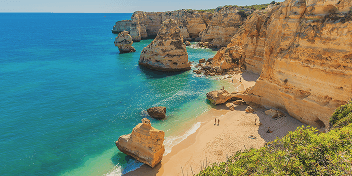 Two people walking on a sandy beach surrounded by rugged orange cliffs and turquoise ocean water.