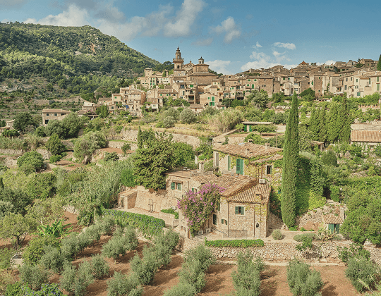 Hilltop village with stone houses, a church tower, and Mediterranean trees under a blue sky.