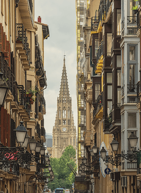 Narrow street lined with tall buildings leading to a Gothic cathedral tower under a cloudy sky.