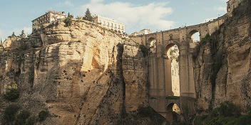 Ancient stone bridge spanning a deep gorge with buildings atop the cliffs in sunny weather.