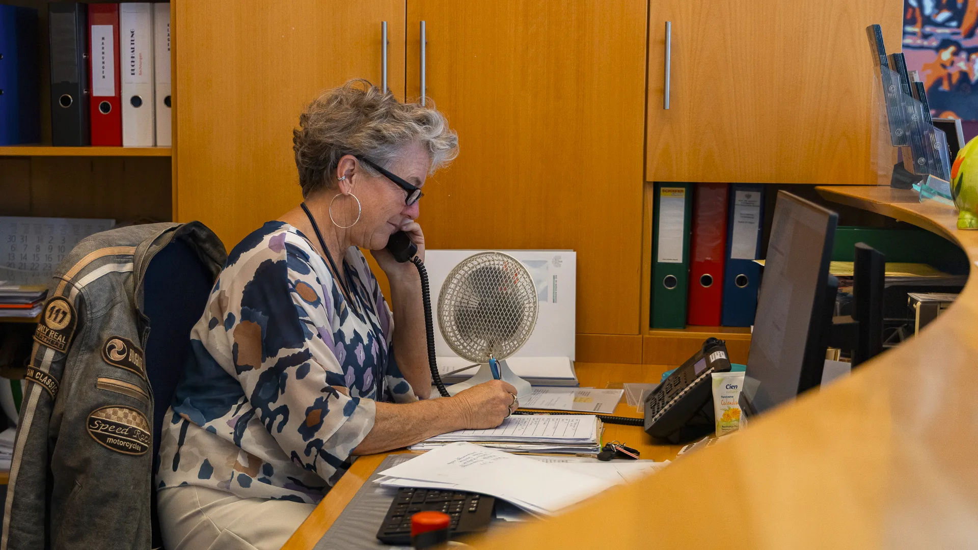 Mitarbeiterin des Notariats Mayrhofer im Empfangsbereich, führt ein Telefonat und macht sich Notizen an ihrem Schreibtisch mit Computer und Aktenordnern.