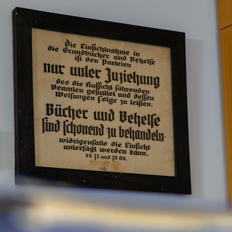 Ein gerahmter, historischer Aushang in Frakturschrift an der Wand der Kanzlei. Der Text erklärt die Regeln zur Einsichtnahme in Grundbücher und mahnt zur schonenden Behandlung der Dokumente.
