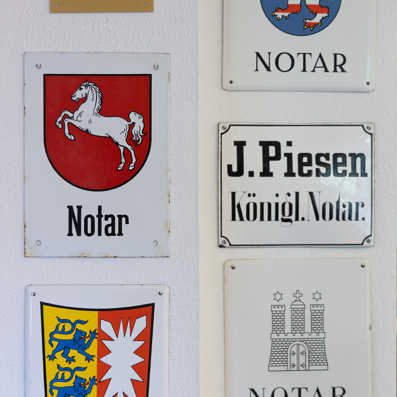 Sammlung verschiedener historischer Amtsschilder in klassischer Typografie für Notare an einer weißen Wand.