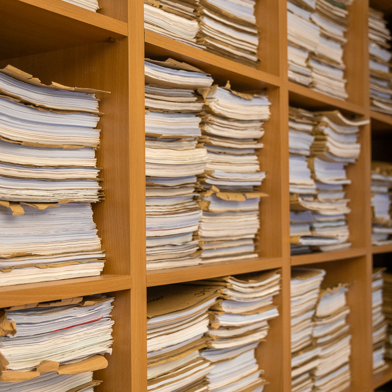 Ein hölzernes Archivregal in schräger Perspektive, gefüllt mit zahlreichen Stapeln aus Papierakten und Dokumenten in braunen Mappen.