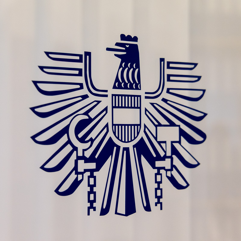 Das offizielle Wappen der Republik Österreich (Bundesadler) als stilisiertes, dunkelblaues Logo, aufgebracht auf einer weißen Säule im Innenbereich der Kanzlei.