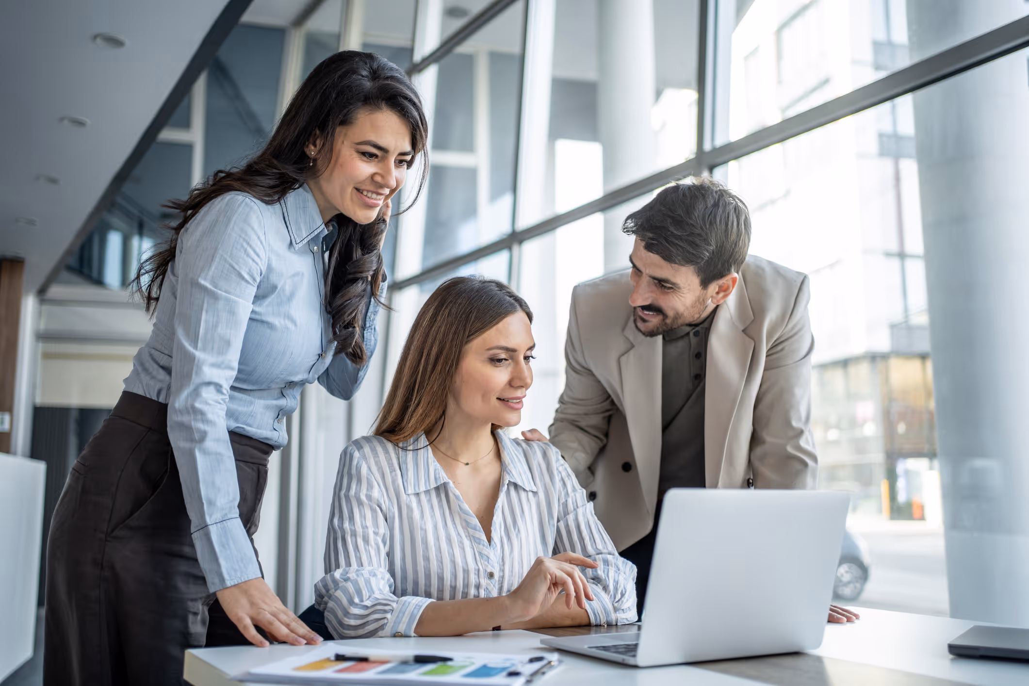 Three colleagues smiling and working together on a laptop in a modern office setting.