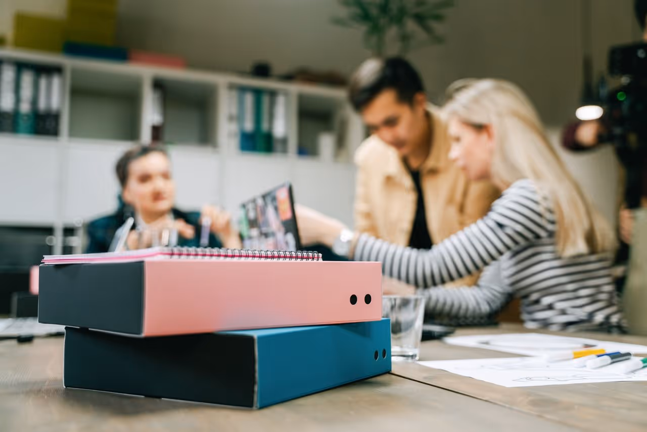 Two ring binders stacked on a table with three people working blurred in background.