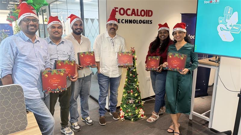 Colleagues in Santa hats holding wrapped gift boxes in front of a Focaloid Technologies wall
