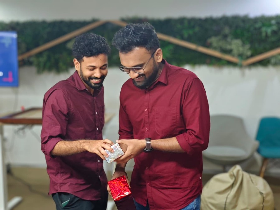 Two colleagues laughing as one opens a wrapped gift in the office