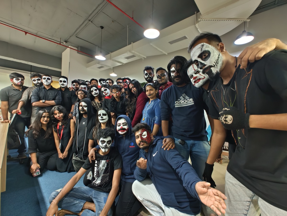 A large group of colleagues in Halloween face paint and black clothing posing together in the office
