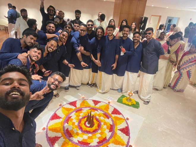 Colleagues in traditional dress around a flower pookalam, one taking a selfie
