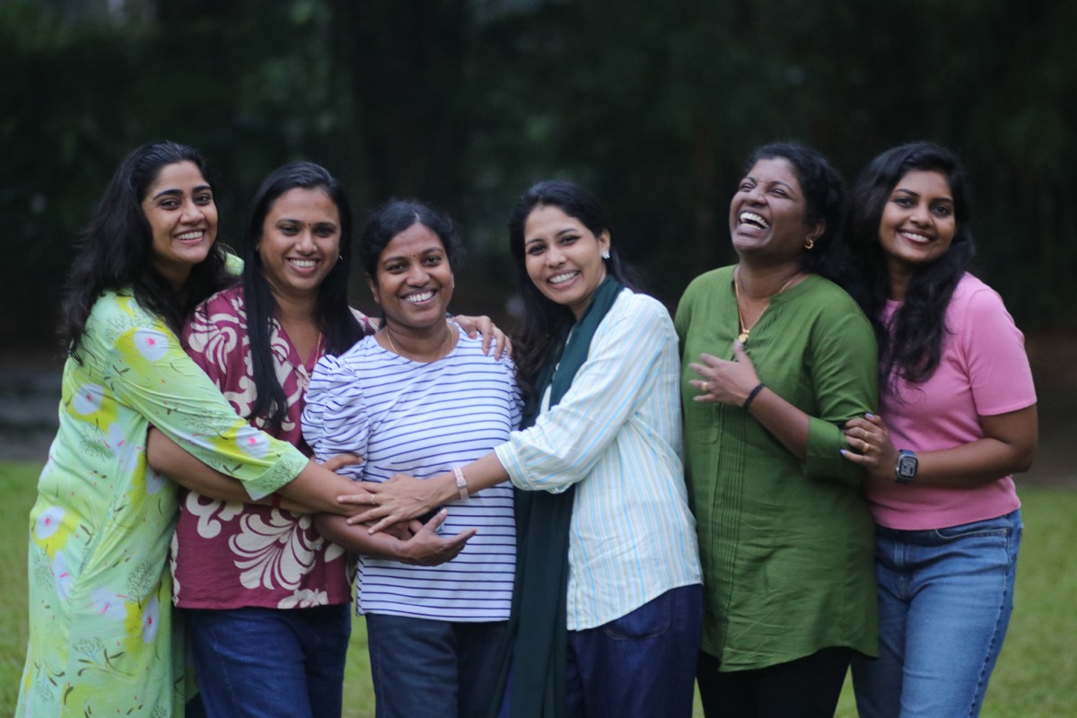 Six colleagues standing arm in arm outdoors, all of them laughing