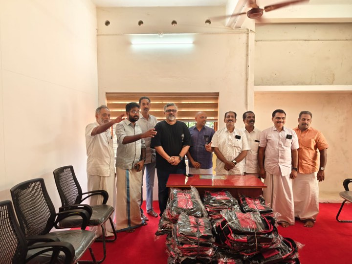 Nine men standing behind a table in a school hall, with a large stack of packaged red and black backpacks on the floor in front of them