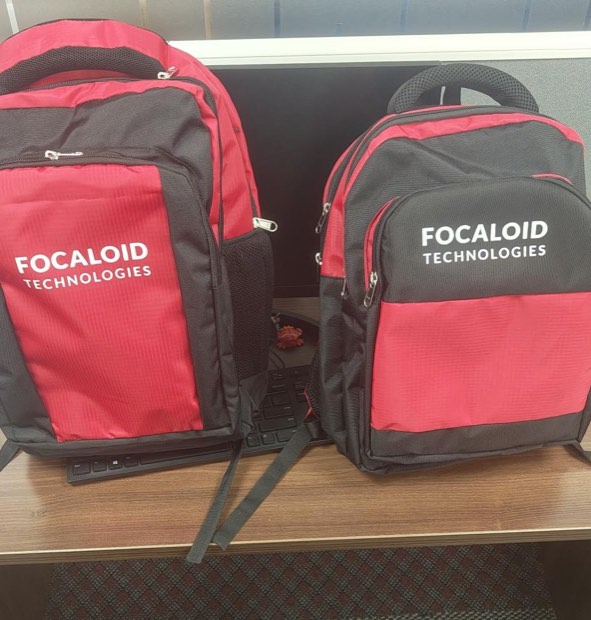 Two red and black backpacks printed with the Focaloid Technologies wordmark, standing on a desk