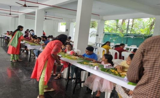 Children and adults seated at long tables being served a meal on banana leaves