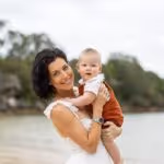 Smiling woman holding a baby near a beach with trees in the background.