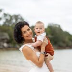 Smiling woman holding a baby near a beach with trees in the background.