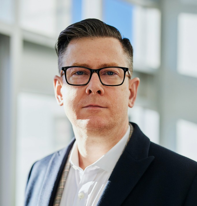 Man with short dark hair wearing black glasses, a white shirt, and a dark blazer standing in a bright indoor setting.